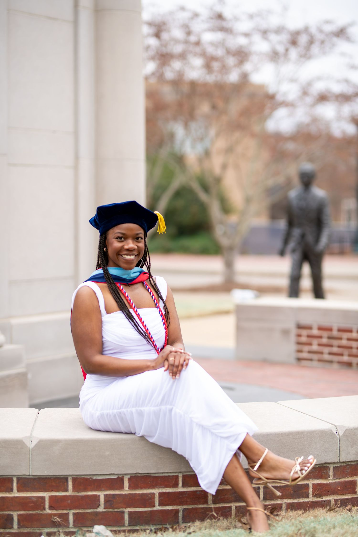 Graduate in academic regalia sits on brick ledge in white dress with graduation cords on college campus.