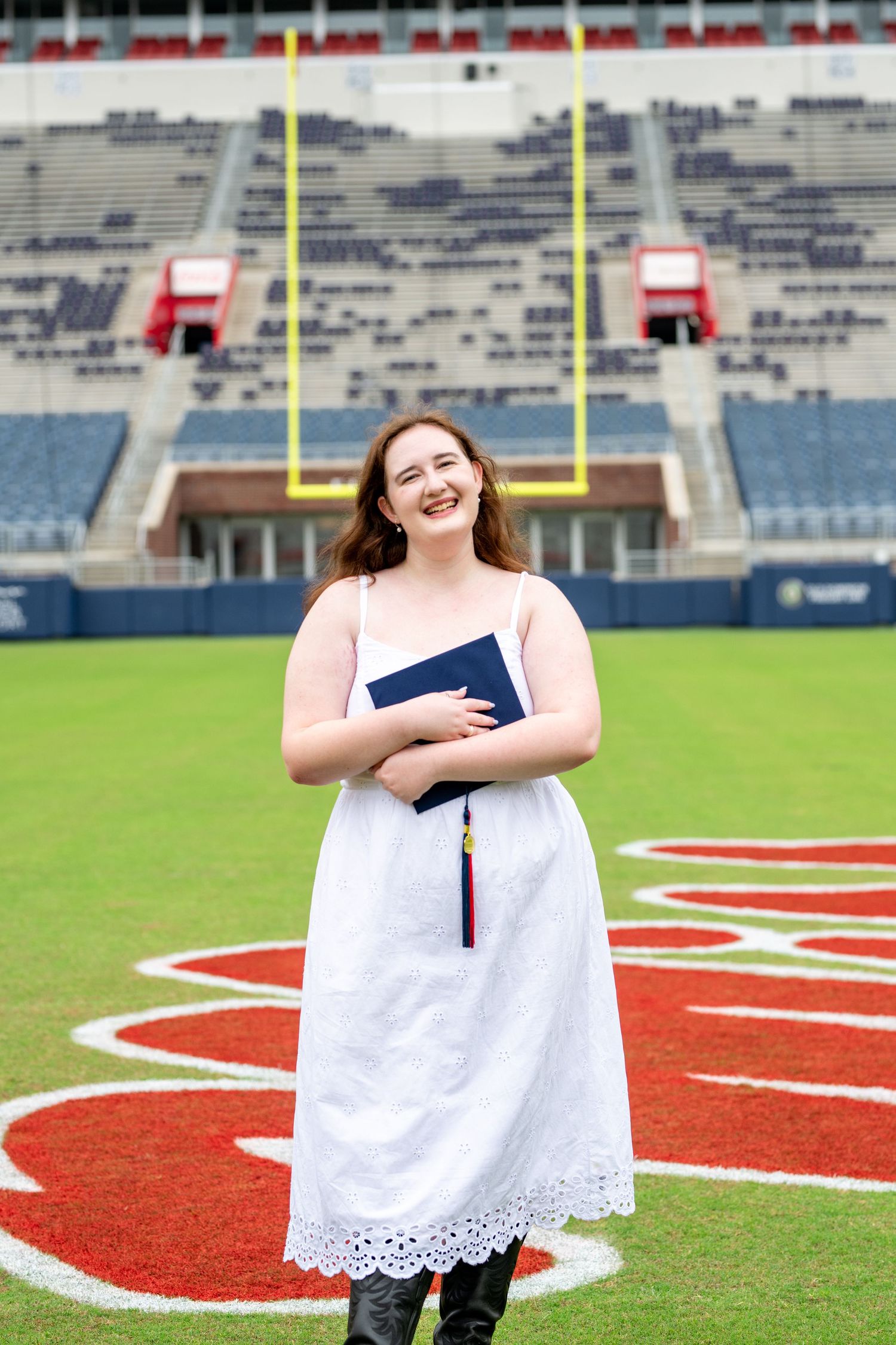 Graduate student poses for commencement photos on a football field with stadium seating and goal posts visible.