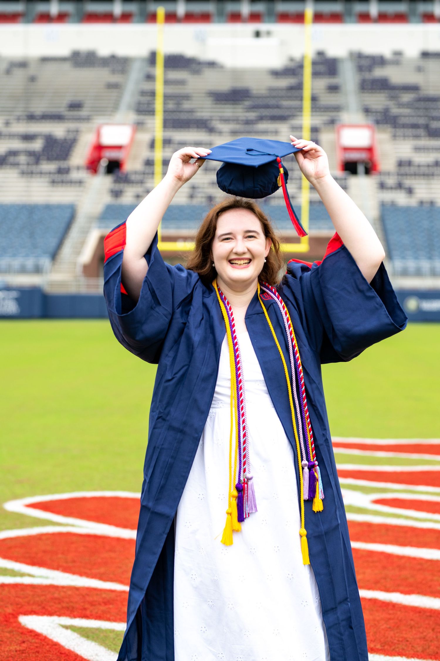A graduate in navy blue cap and gown celebrates on a football field with stadium seating in the background.