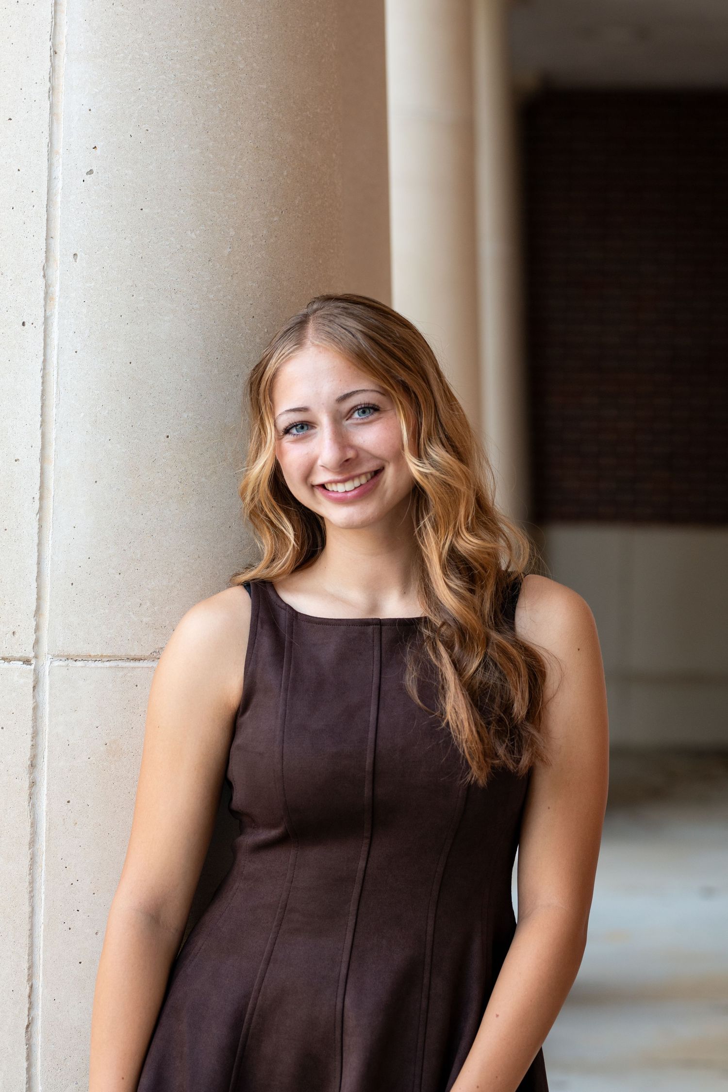 Young woman with long blonde hair smiling in a brown sleeveless dress, posing beside stone columns outdoors.