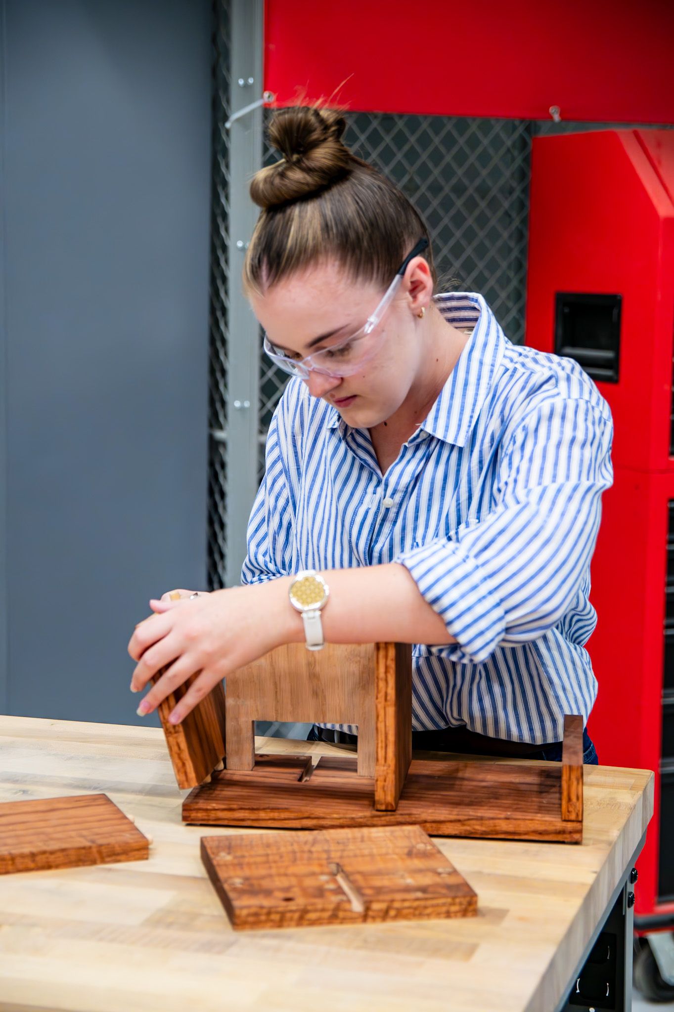 Young woman wearing safety glasses assembles a wooden box project on a workbench in a workshop.
