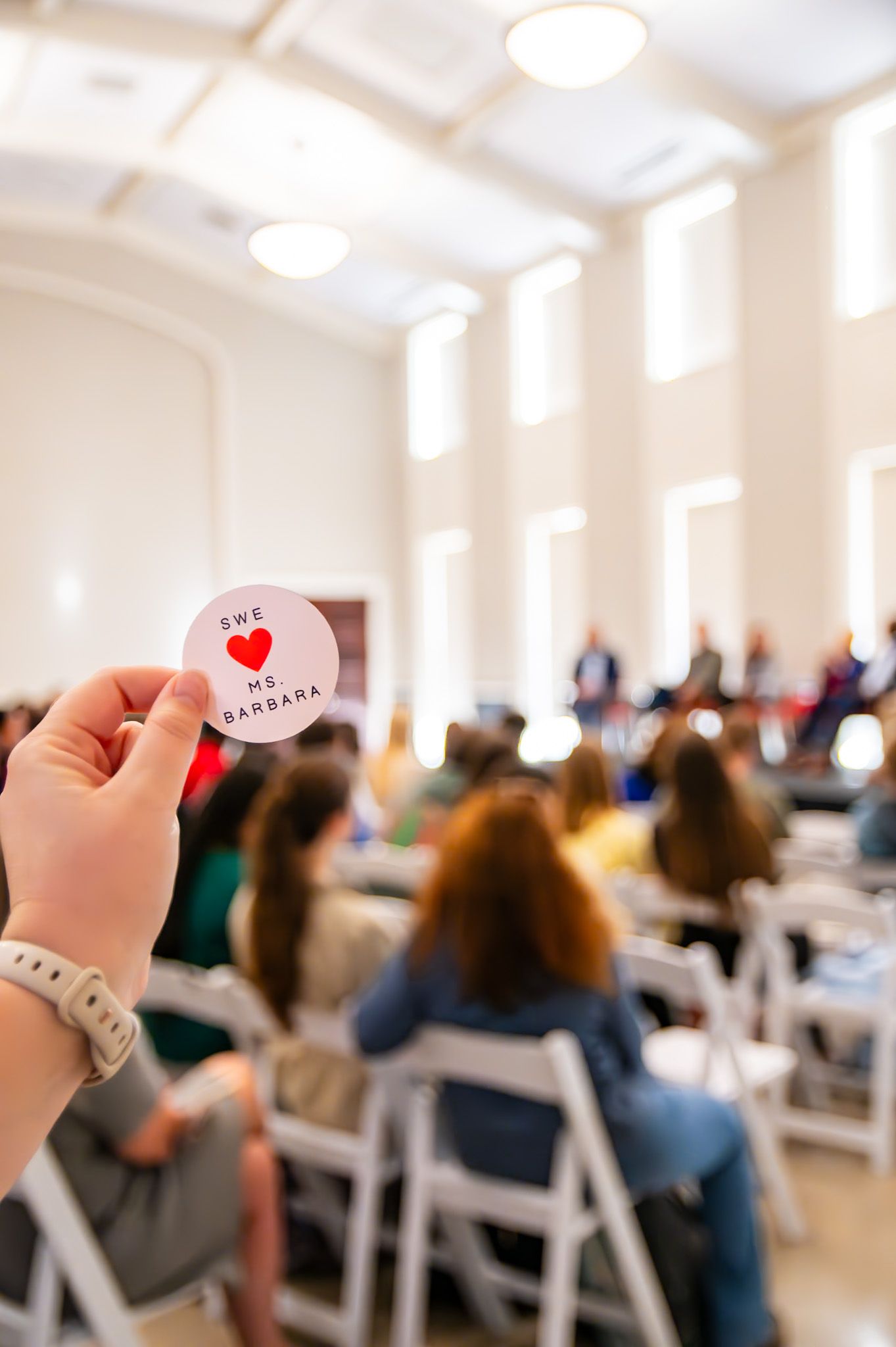 Hand holding a button reading 'We Love Ms Barbara' at an indoor event with attendees seated in white chairs.