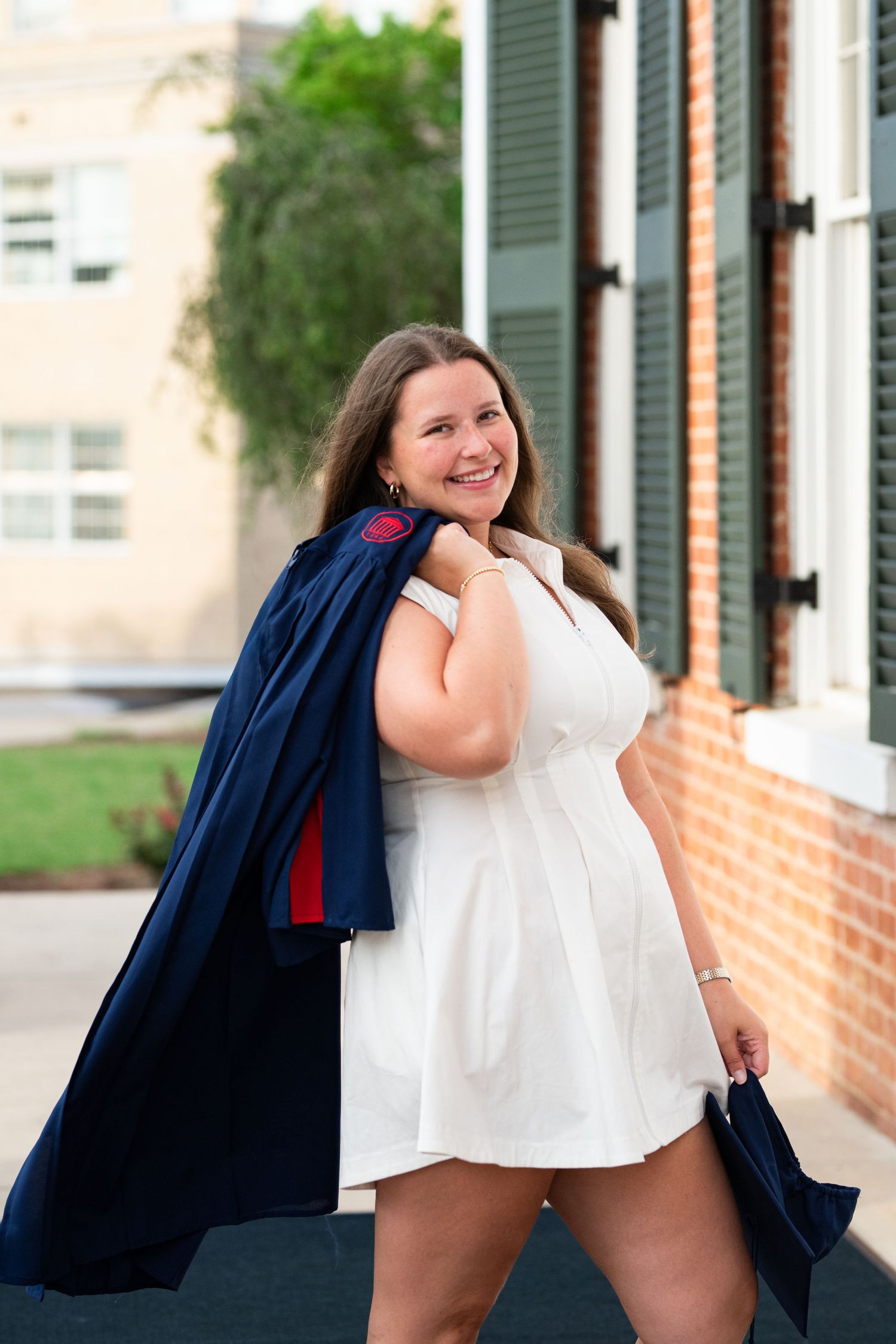 Smiling young woman in white dress holding navy blue graduation gown over shoulder outdoors near brick building.