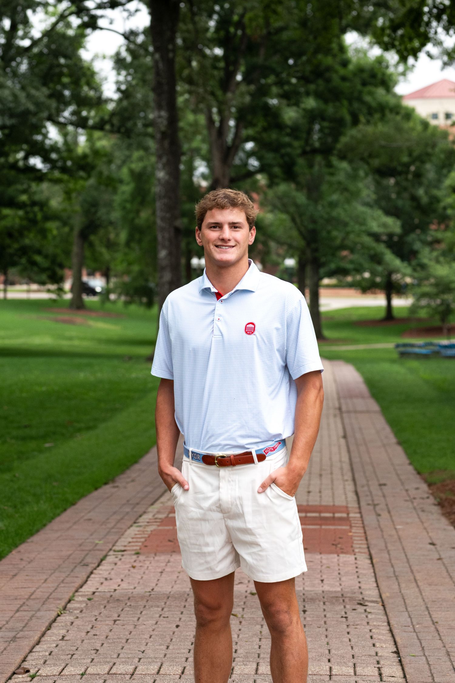 Young man in light blue polo and white shorts smiling on a brick pathway surrounded by green trees.