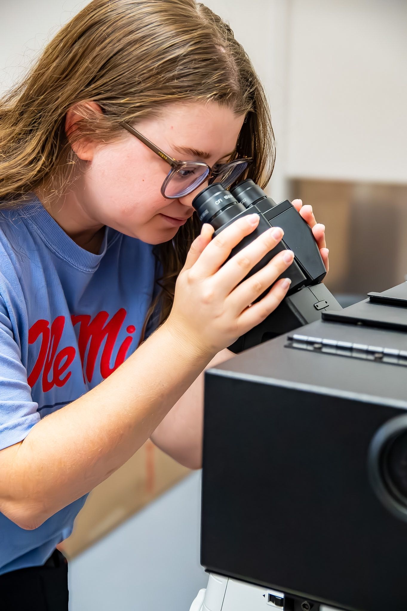 A young woman wearing glasses and an Ole Miss t-shirt looks through a microscope in a laboratory setting.