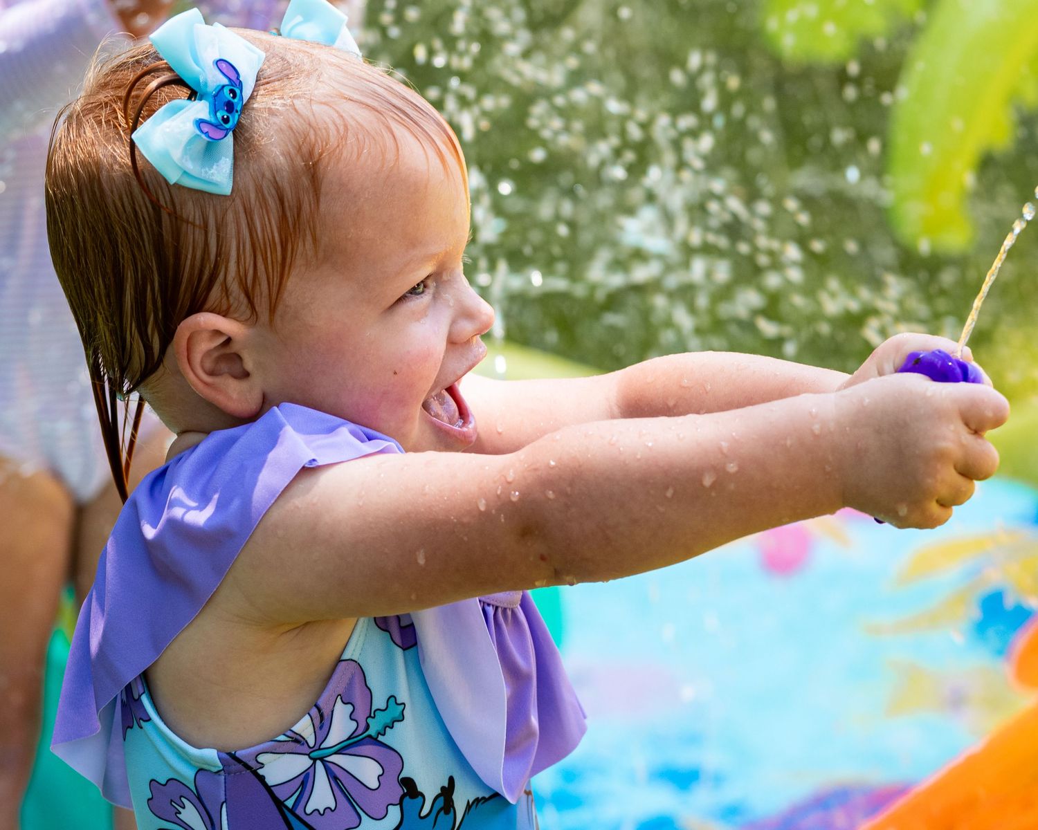 Toddler girl with blue bow squirts water gun at outdoor pool party in colorful swimsuit.