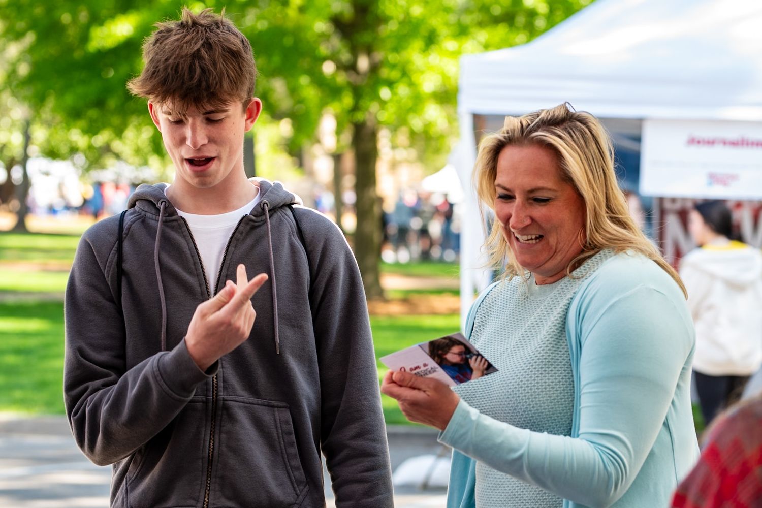 Teen boy and woman smiling at outdoor market event, boy gesturing while woman holds smartphone.