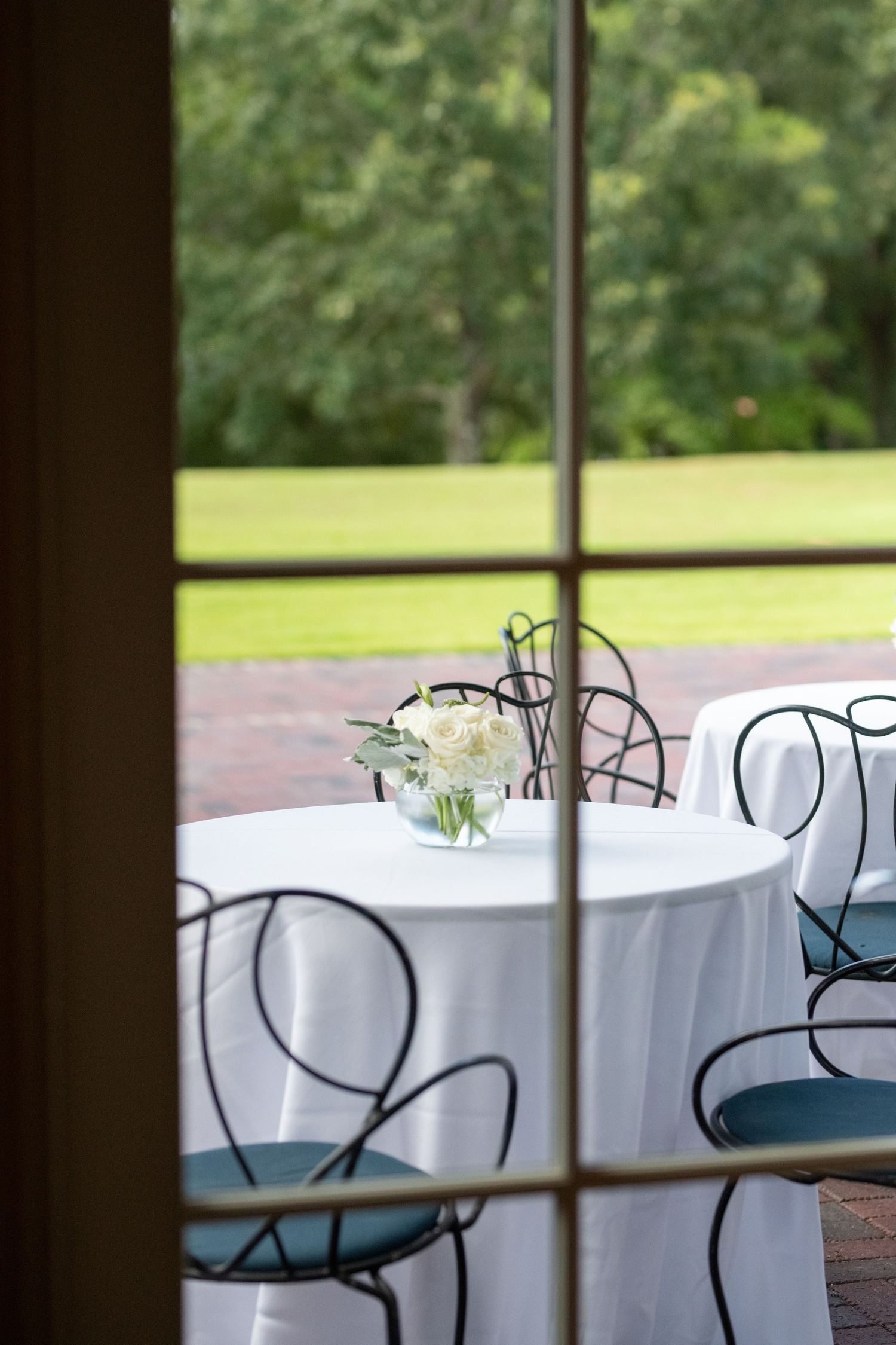 White tablecloth covered table with blue chairs seen through window overlooking garden setting.