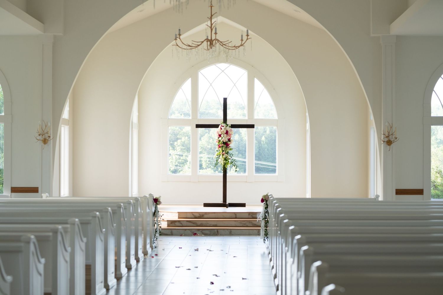 Light streams through arched windows in a pristine white church sanctuary with rows of empty wooden pews.