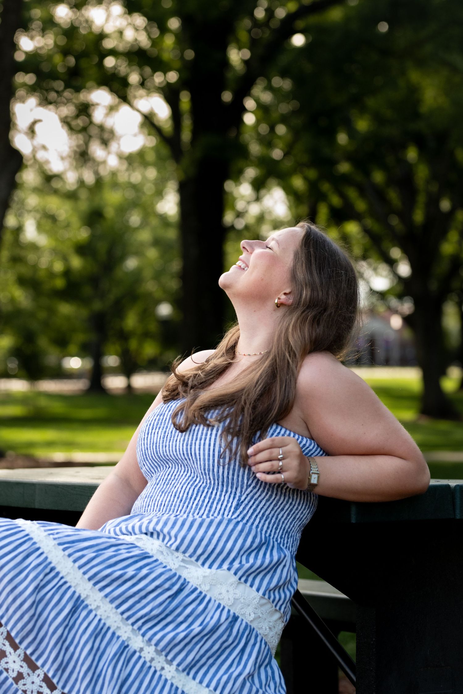 Woman in blue striped sundress laughing joyfully on a park bench surrounded by lush green trees.