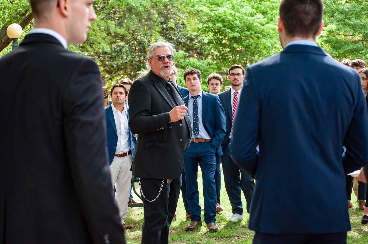 Guests in formal navy suits gather outdoors for a social event in a garden setting.