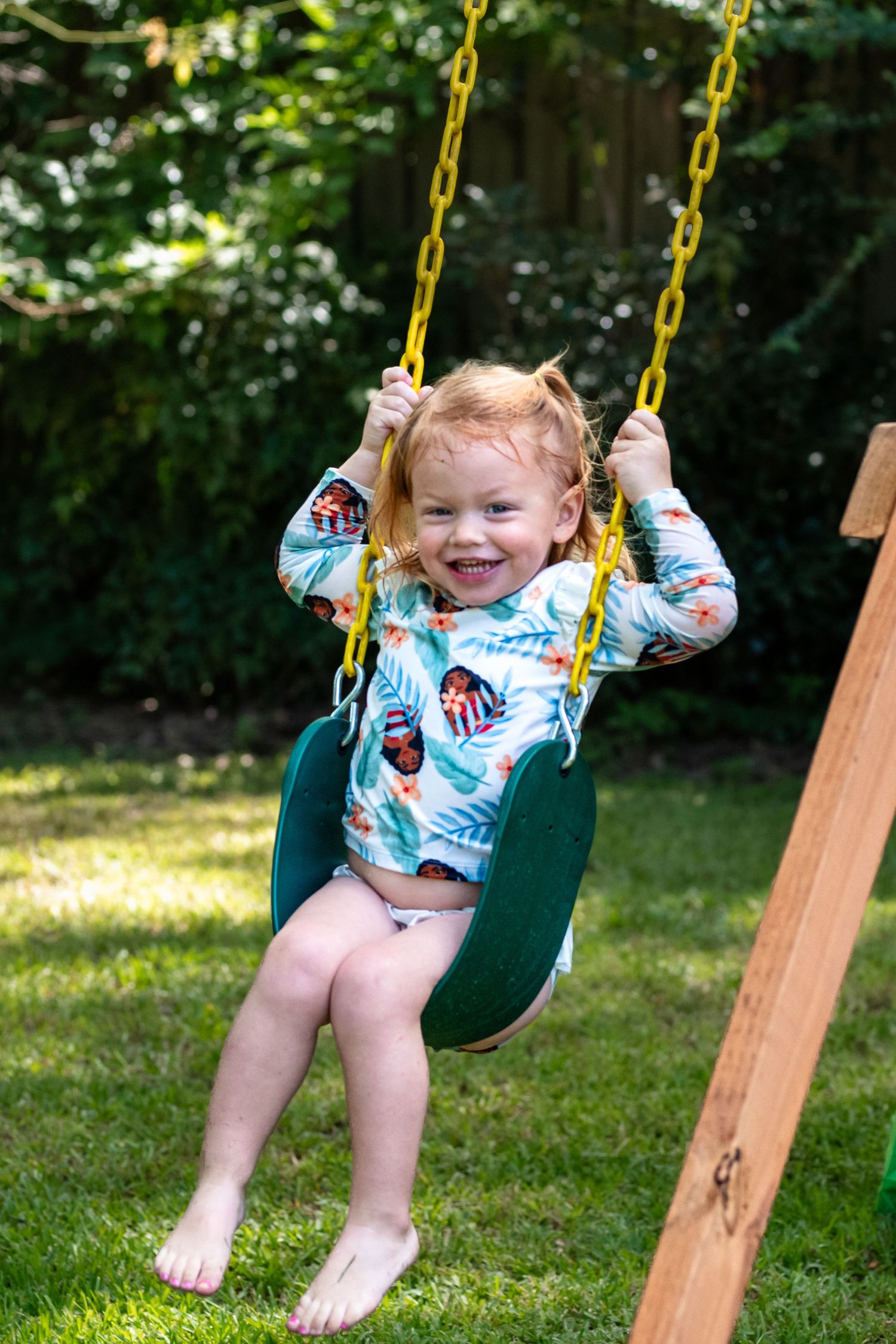Smiling toddler girl with red hair sitting on a green swing with yellow chains in a sunny backyard.