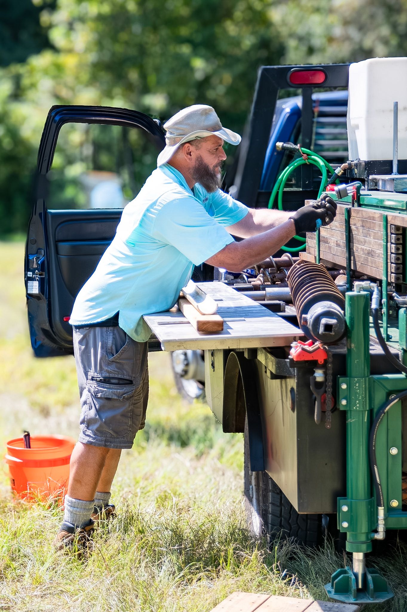 A bearded man in a light blue polo and cap operates equipment on a work truck outdoors on a sunny day.