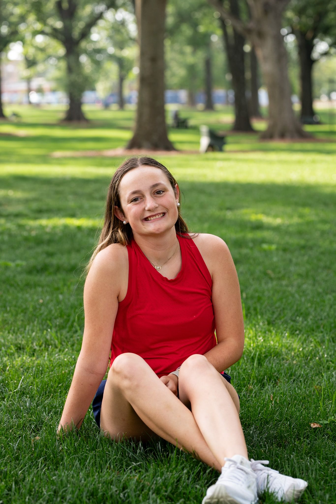 Young woman in red tank top sitting cross-legged on green grass in a park, smiling cheerfully.
