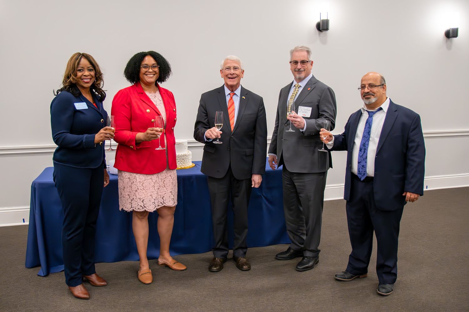 Five professionals pose together smiling at a formal networking event with a blue draped table in the background.