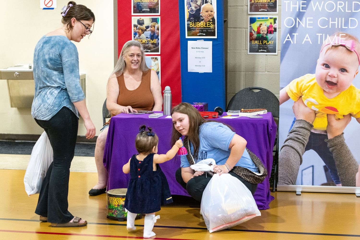 Women and toddler at a purple-draped booth in a gymnasium with child charity banners displayed.