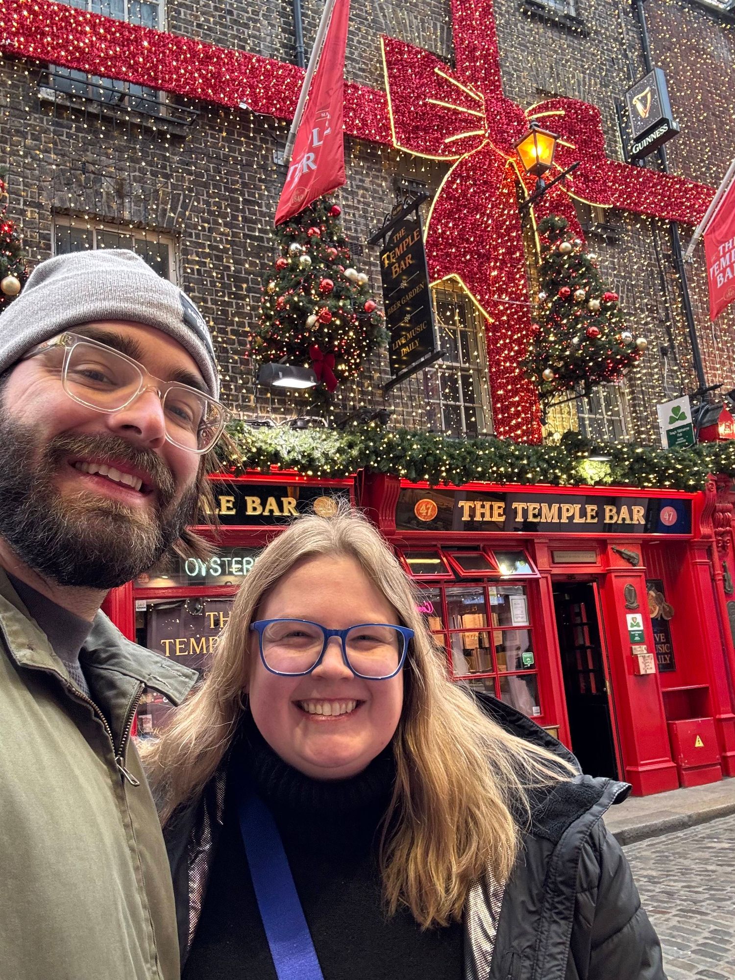 Couple smiling in front of The Temple Bar pub in Dublin, Ireland, decorated with Christmas lights and a giant red bow.