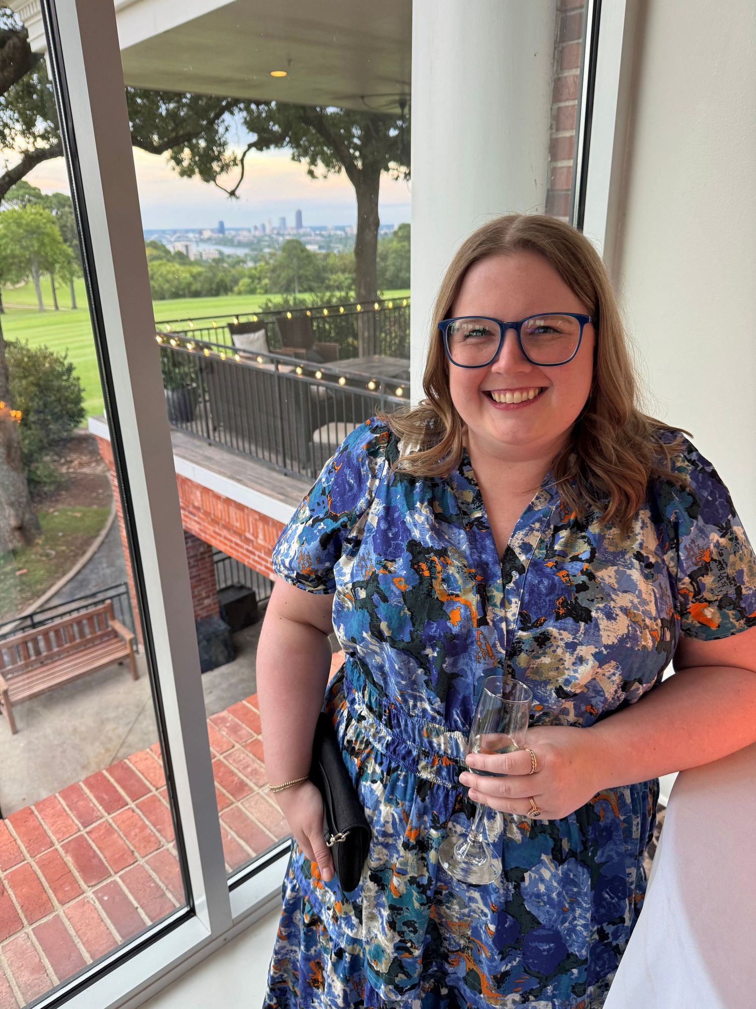Woman in blue floral dress smiling on a balcony with scenic city skyline view in background.