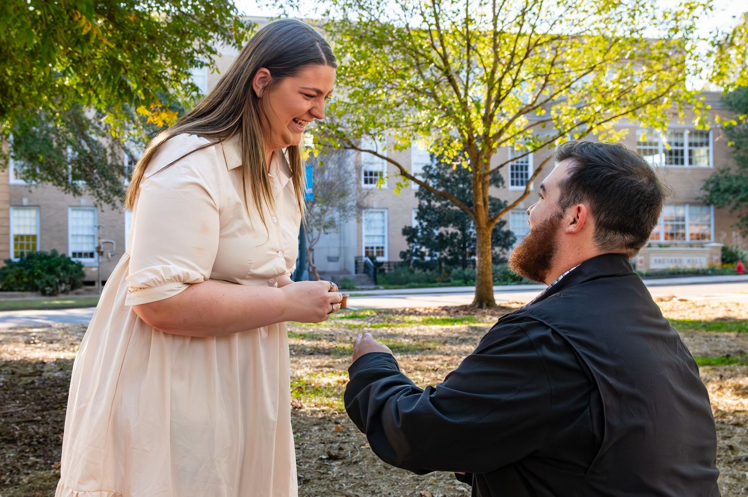 A person kneels on one knee while proposing in a park on a sunny autumn day as trees sway in the background.