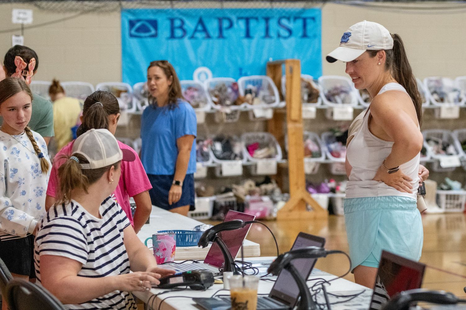 Volunteers assist community members at a Baptist charity shoe drive event indoors with tables of shoes displayed in background.