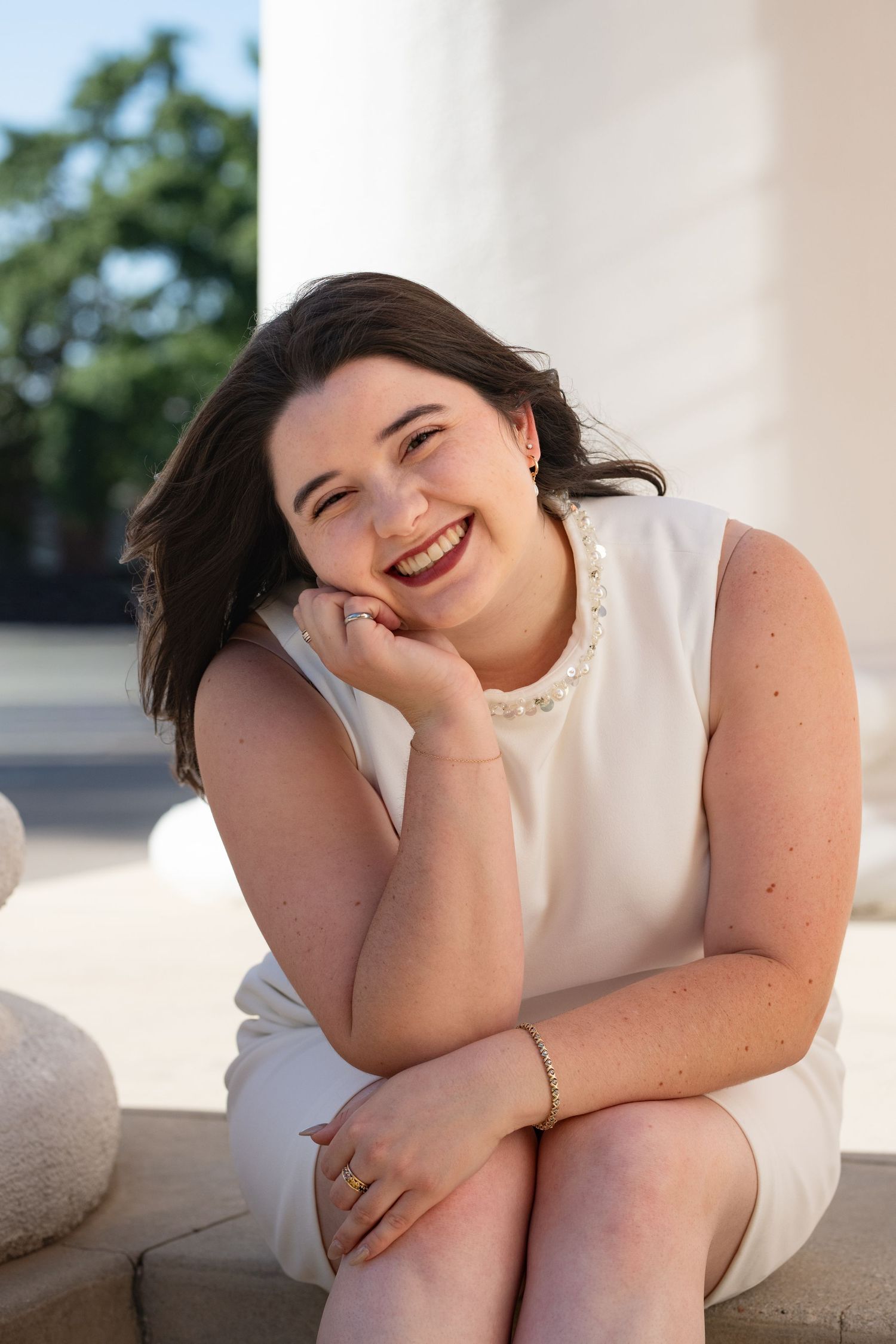 Smiling young woman in white sleeveless dress sitting on steps near columns, wearing pearl jewelry.