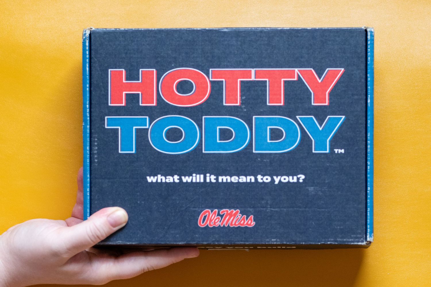 Hand holding an Ole Miss Hotty Toddy branded navy blue gift box on a yellow background.