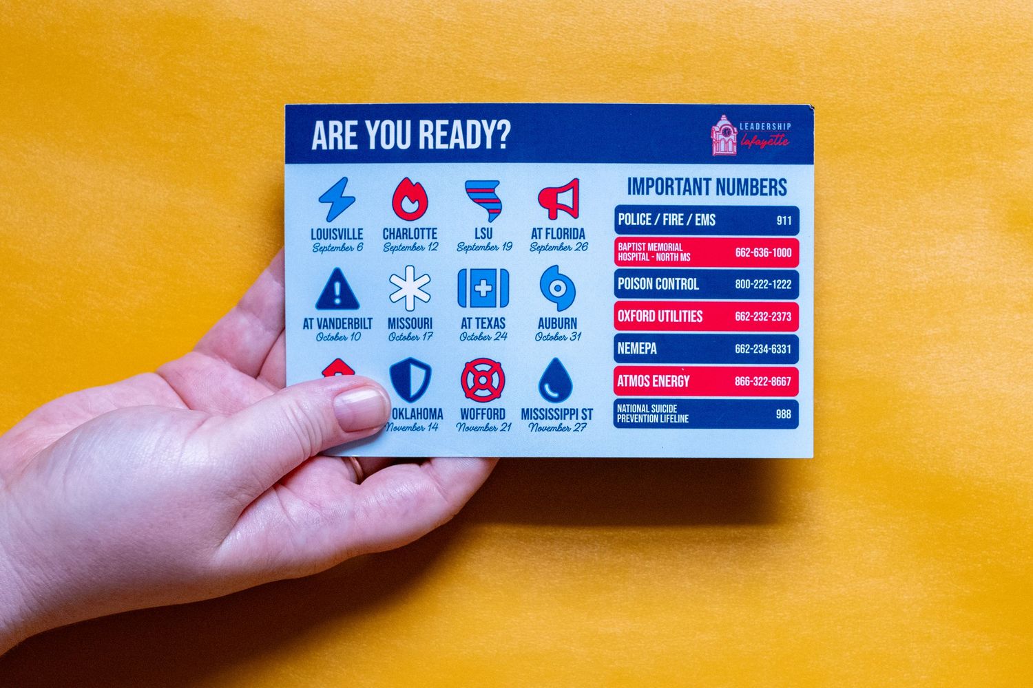 Hand holding an emergency preparedness card with important numbers and safety icons on a yellow background.