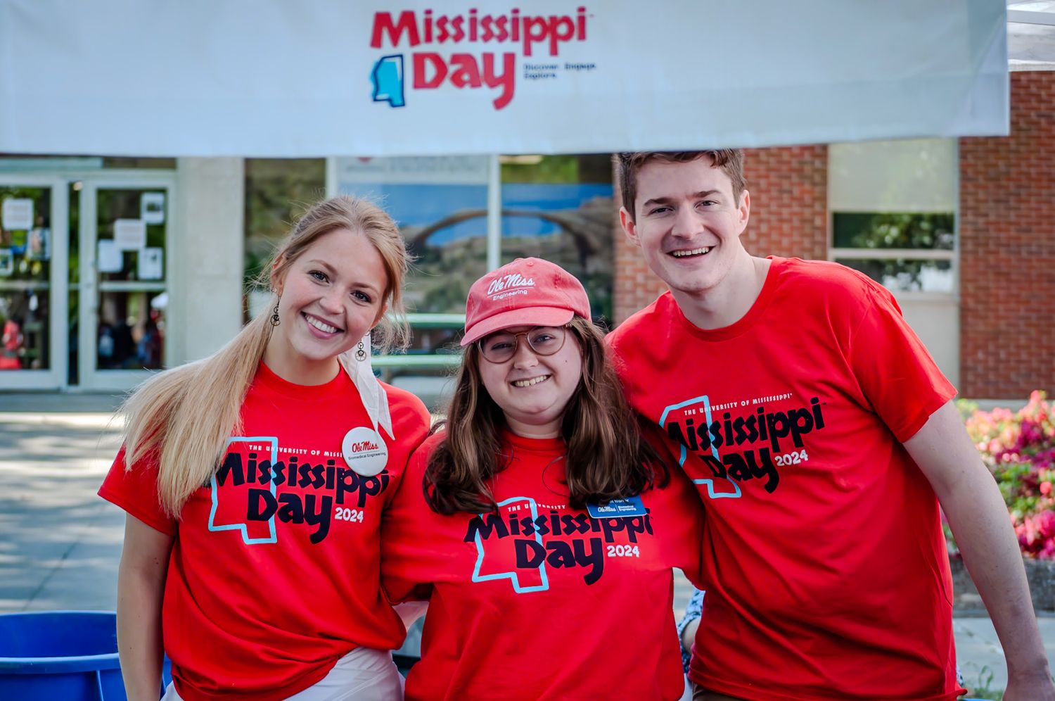 Three volunteers in matching red Mississippi Day t-shirts smile together at an outdoor event.