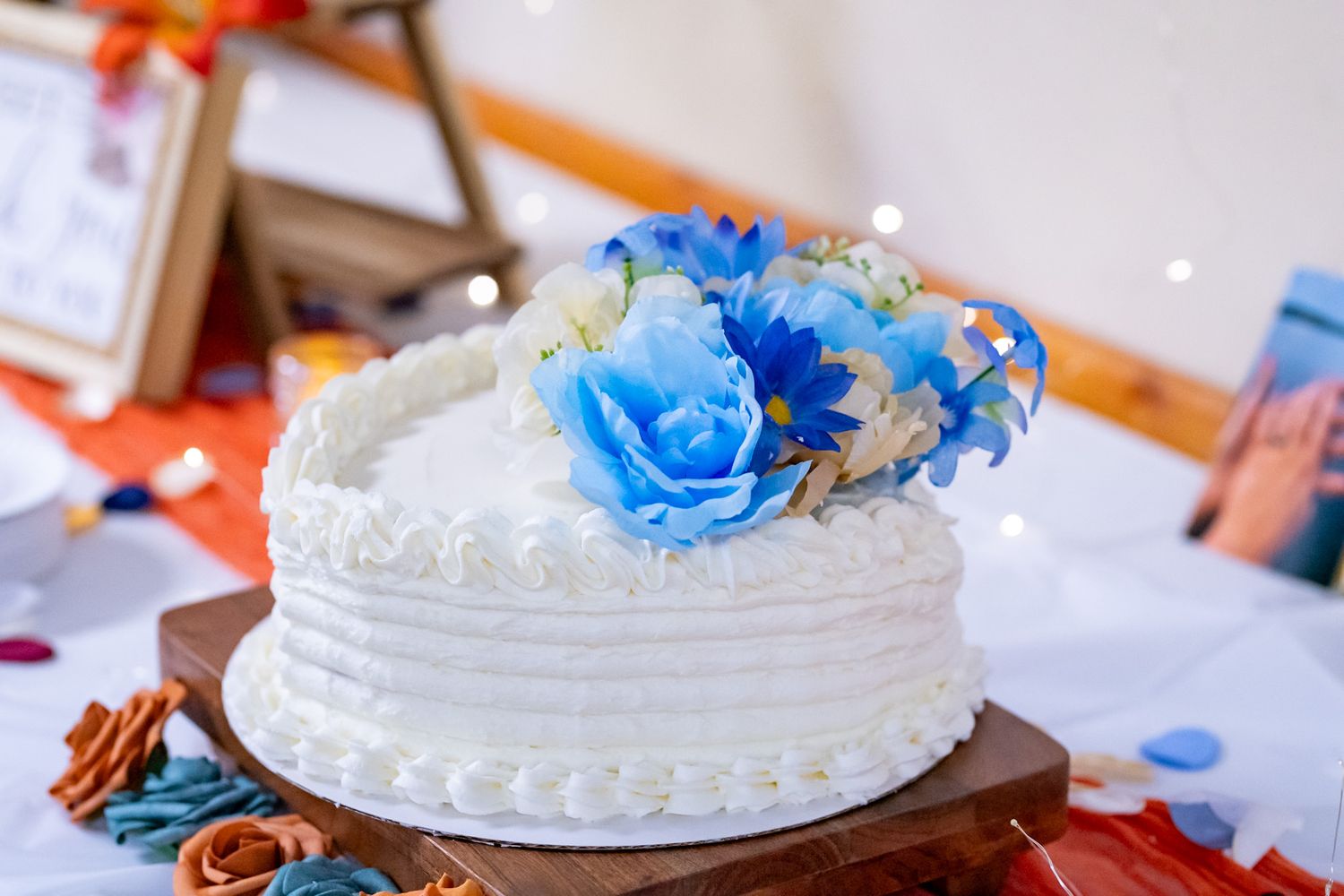 A wedding cake decorated with white frosting and blue roses sits on a decorated table at a reception.