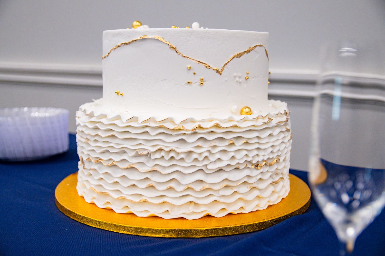 White two-tier wedding cake with ruffled layers and gold accents displayed on a gold base against a blue tablecloth.