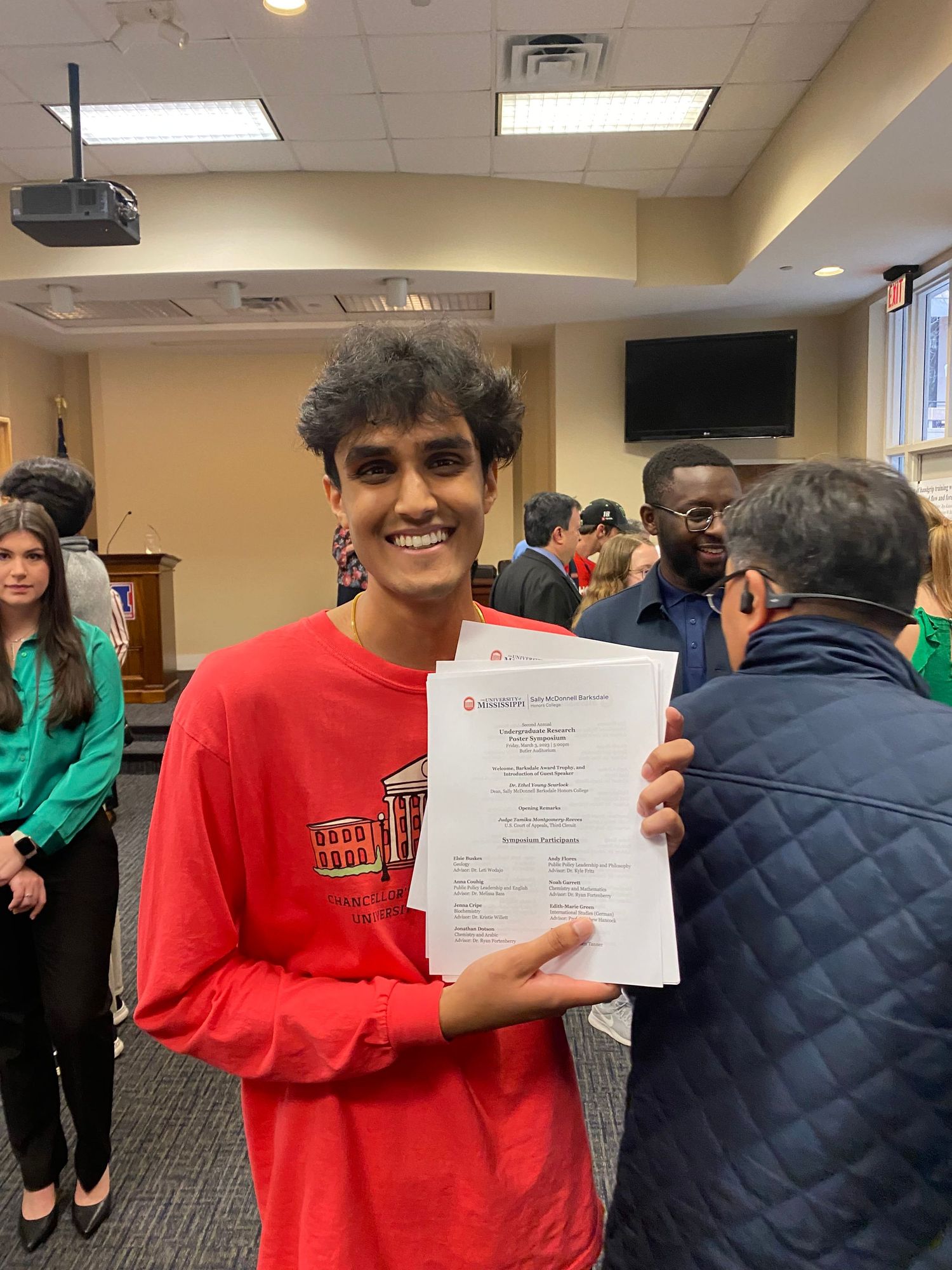 Smiling young man in red shirt proudly holding certificates at an indoor academic awards ceremony.