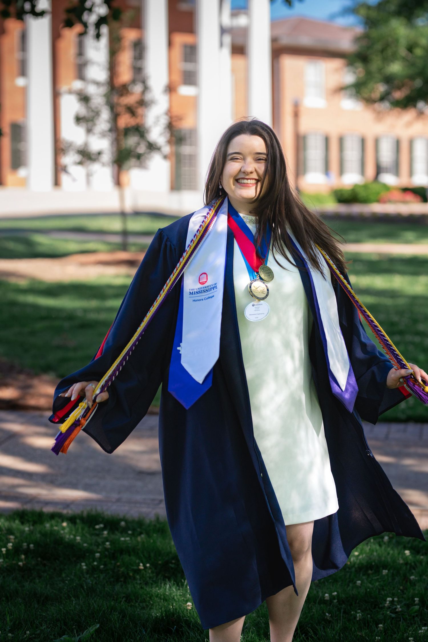 Smiling female graduate in navy cap and gown with gold medal and honor cords poses on college campus.