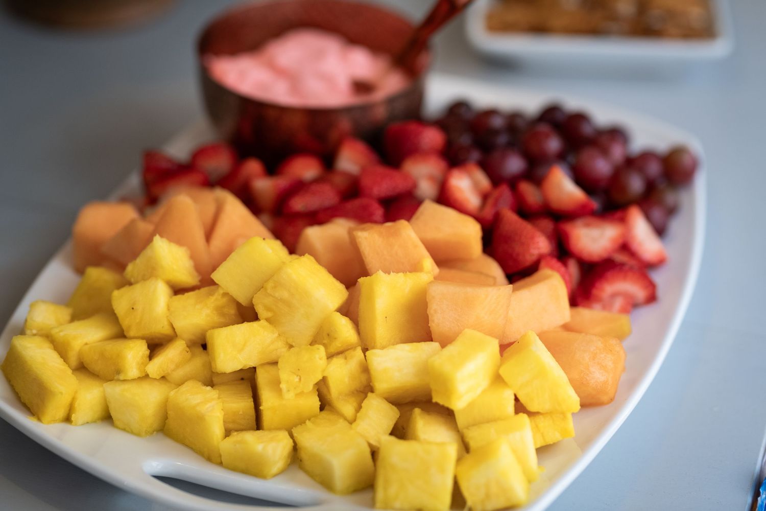 Fresh fruit platter with pineapple, cantaloupe, strawberries, grapes, and pink dipping sauce on a white plate.