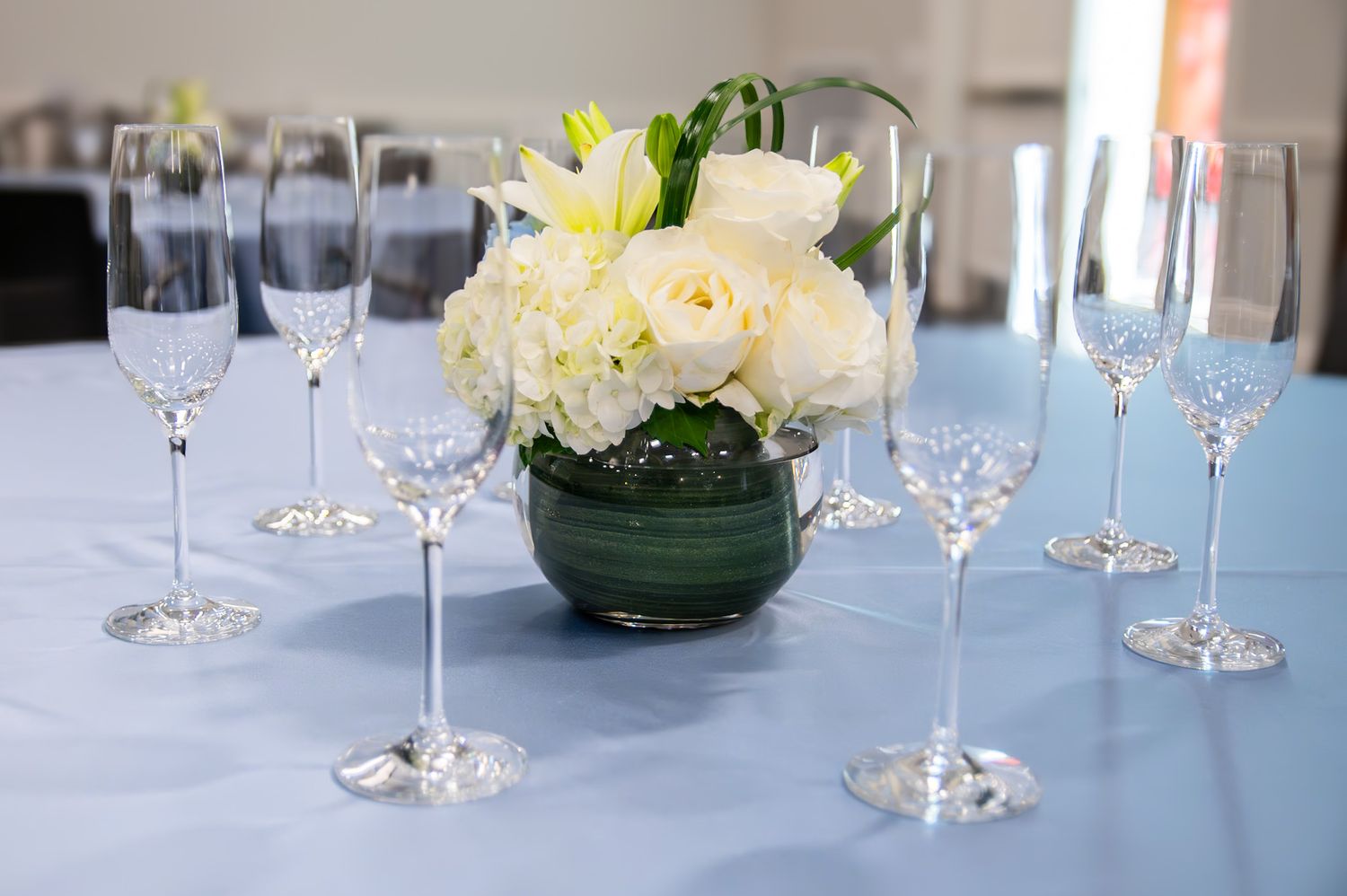 Crystal champagne flutes arranged around a white rose centerpiece in a green vase on a light blue tablecloth.