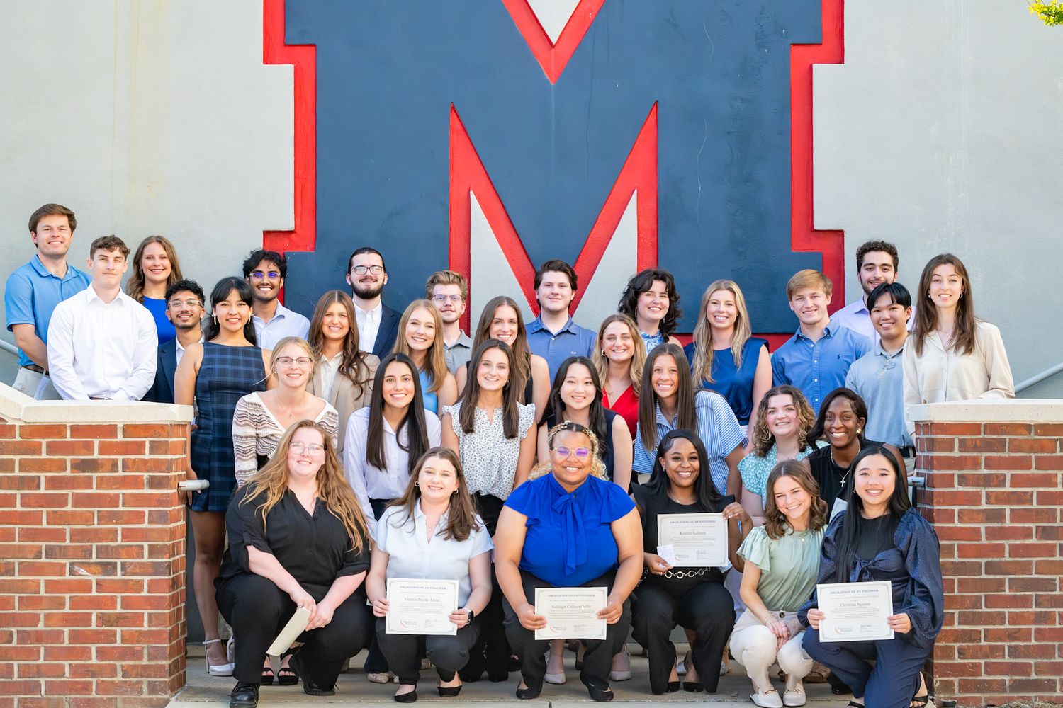 Large group of people stand together holding certificates in front of a red brick building wall.