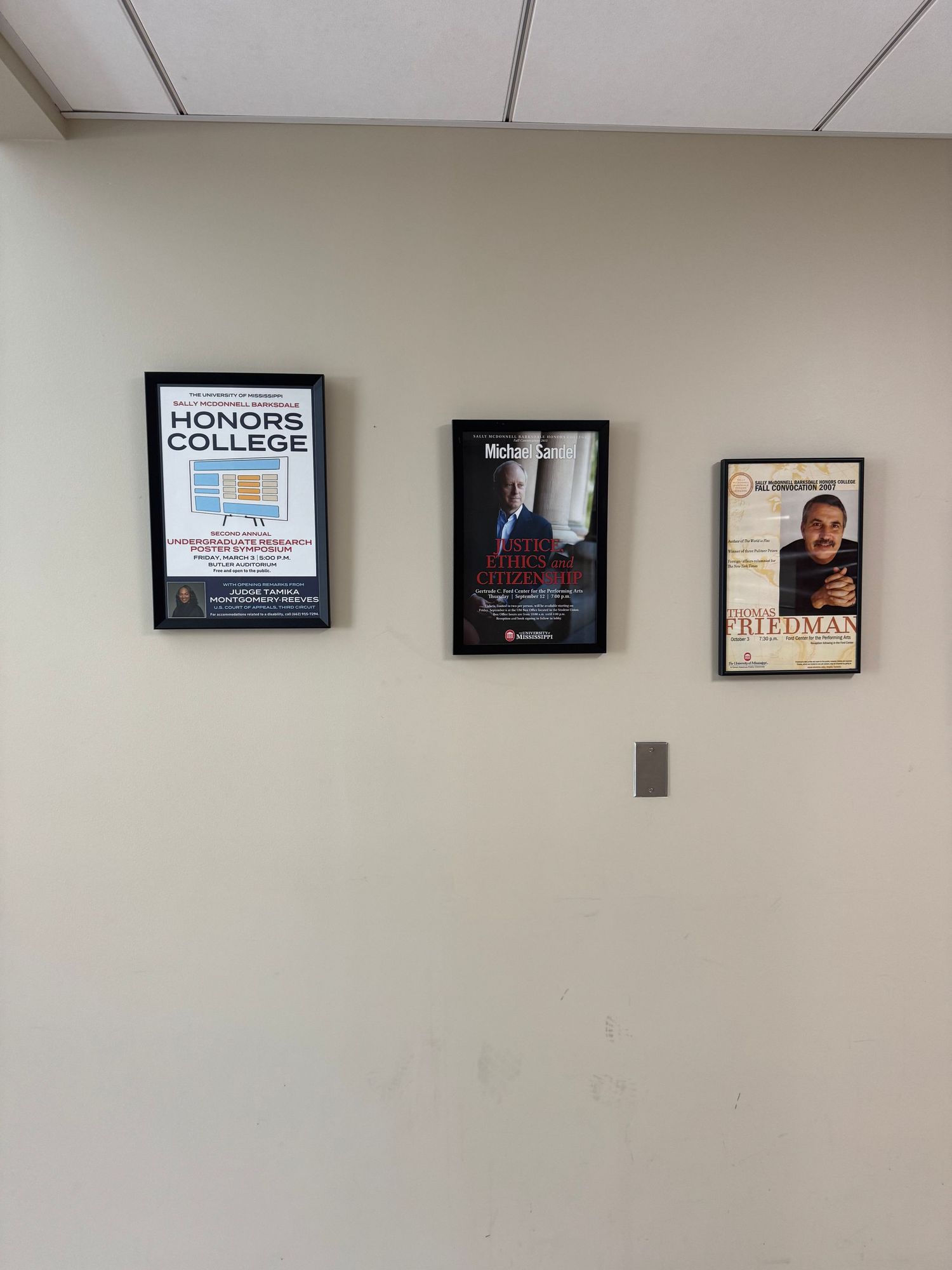 Three framed posters on a beige wall featuring Honors College, Michael Moore, and Milton Friedman educational displays.