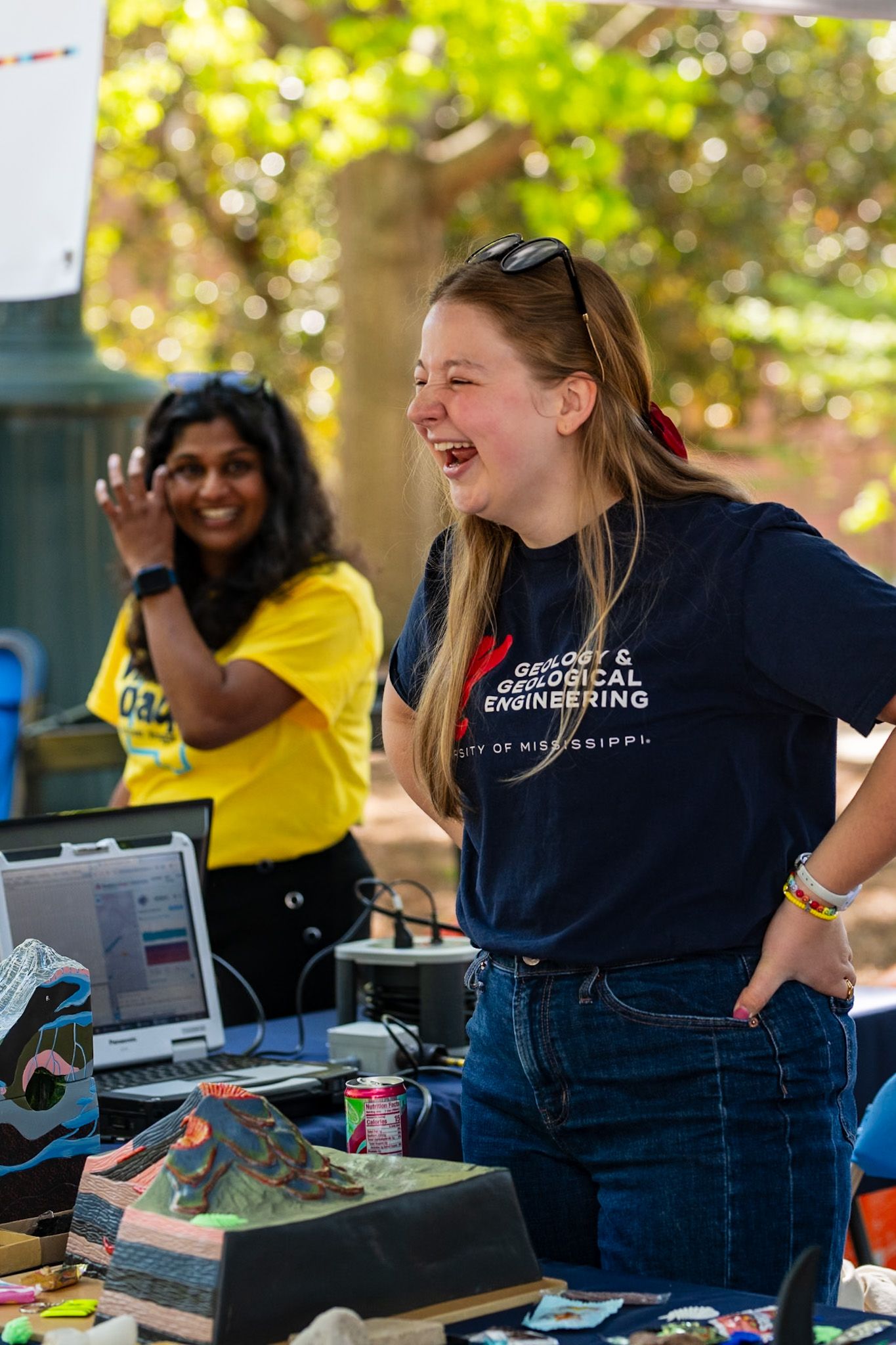 Joyful students laugh together while staffing an information table outdoors with electronics and materials on display.