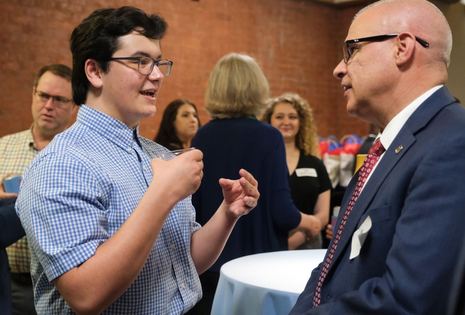 Two men engaged in conversation at a networking event, with guests socializing in the background.