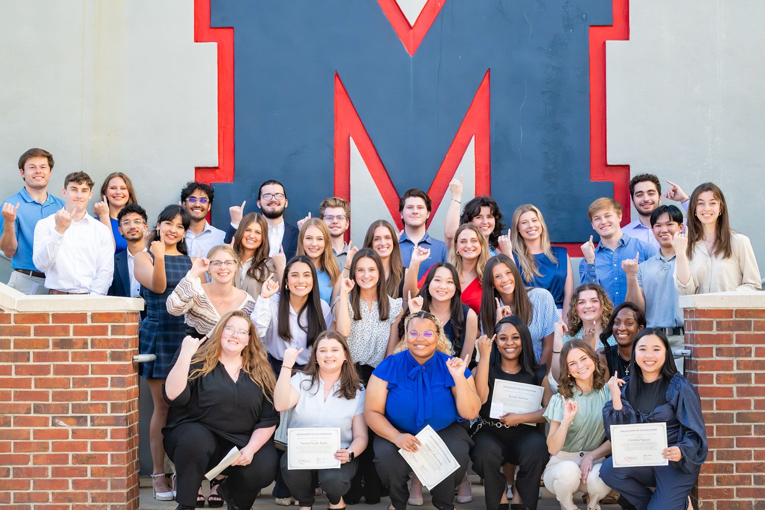 Group of university students posing proudly in front of a large red M logo on a brick wall, holding certificates.