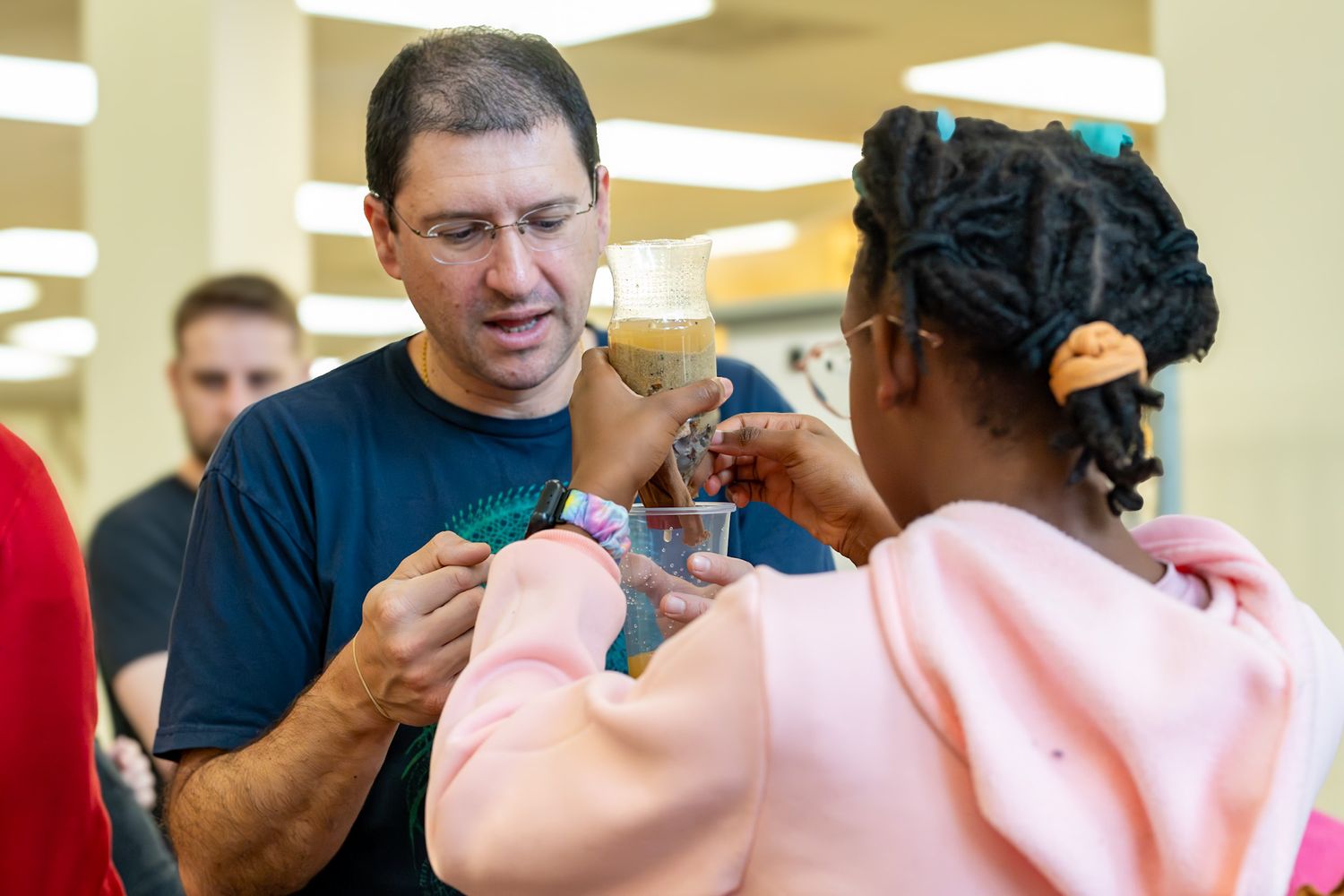 A man in a blue shirt interacts with a young girl holding a cup at an indoor educational event.