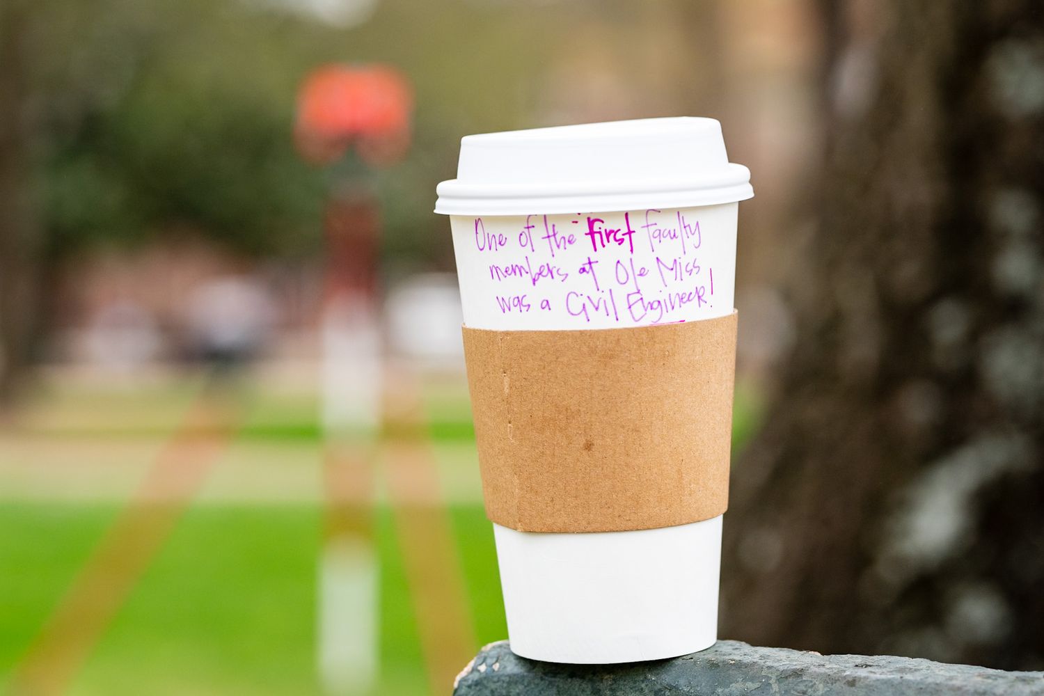 A to-go coffee cup with a cork sleeve and handwritten message displayed outdoors on a stone surface.