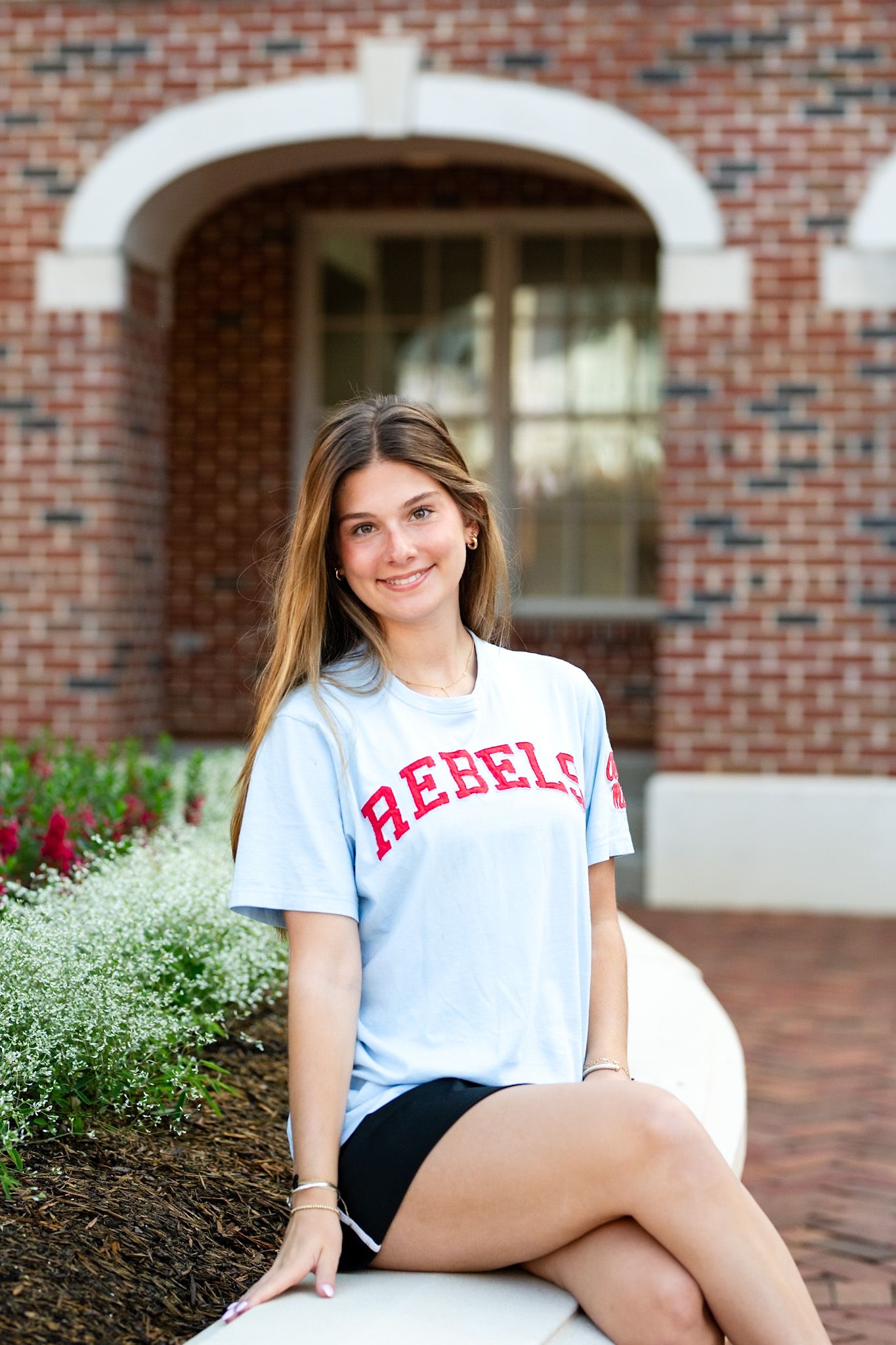 Young woman wearing a Rebels t-shirt sits smiling outdoors near a brick building with flowers.