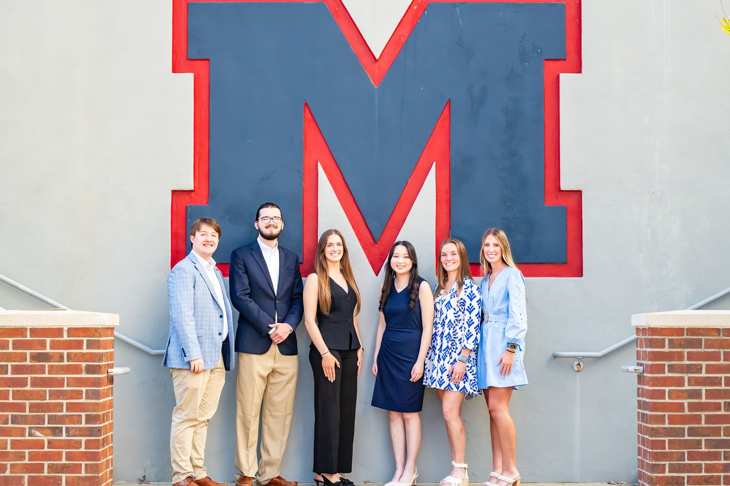 Six students pose together in front of a large red and blue letter M on a gray wall at a school event.
