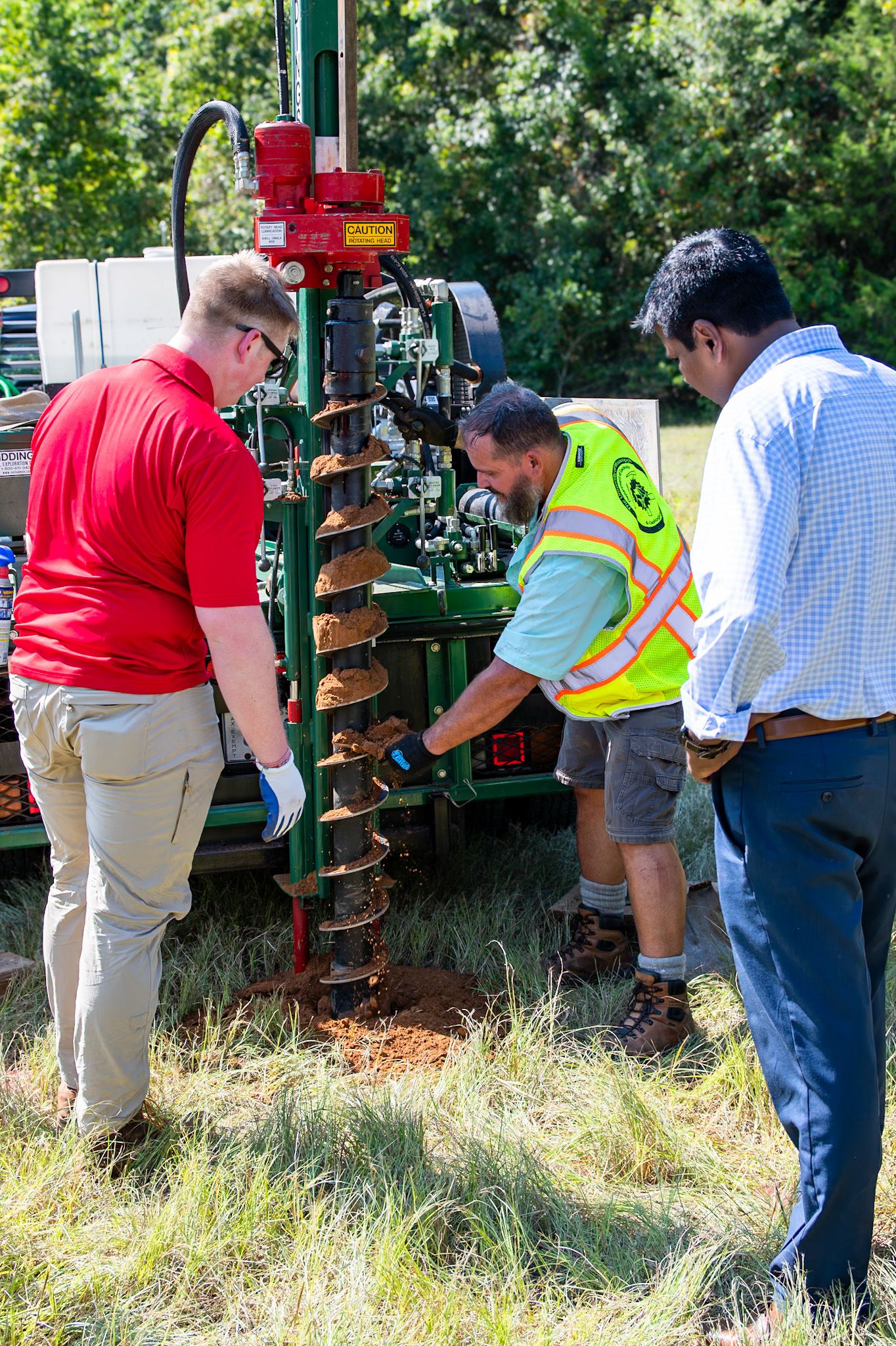 Three workers inspect a green auger drilling machine outdoors, one wearing a safety vest, on a grassy field.