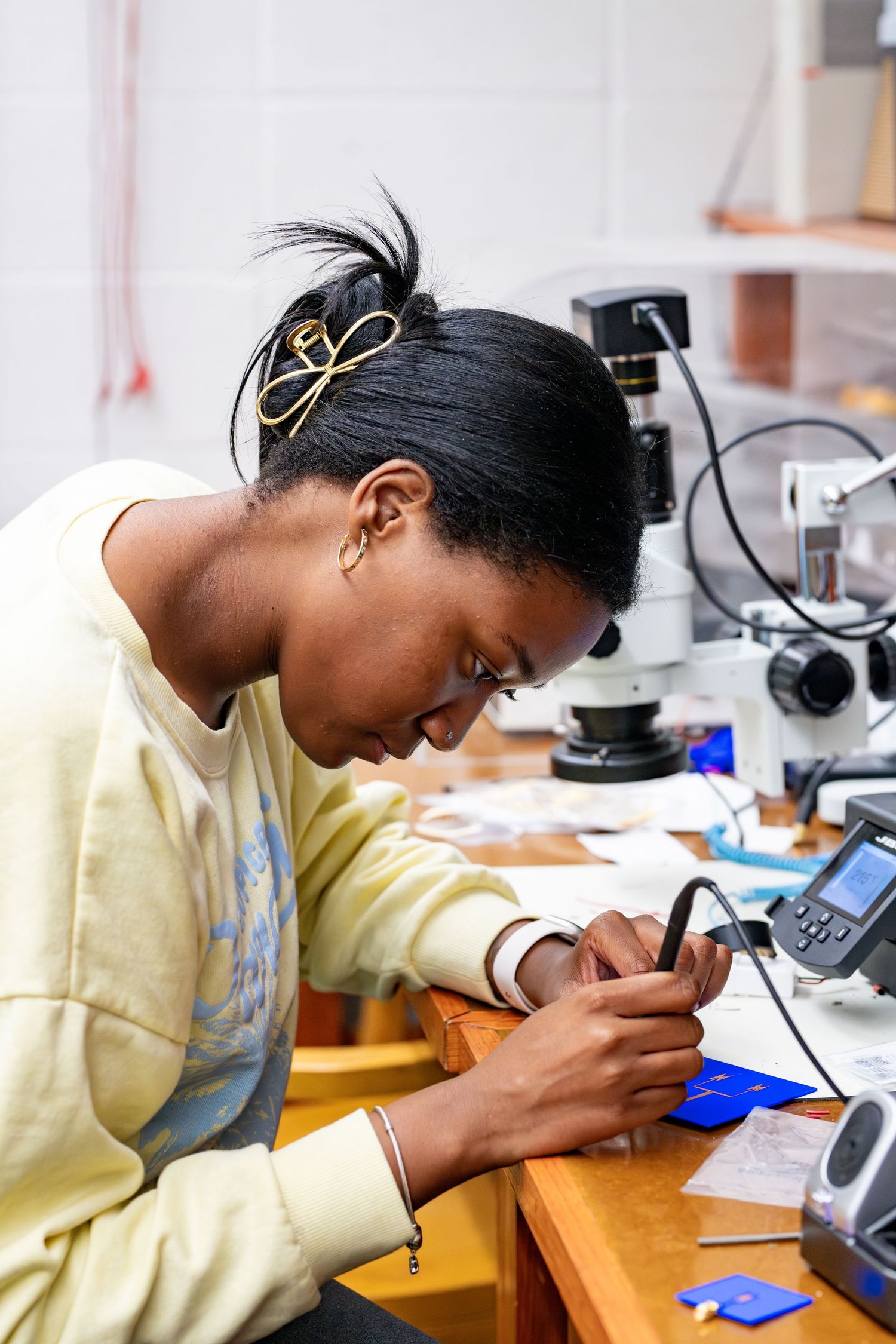 A young Black woman in a lab carefully works with precision tools at a workbench beside a microscope.