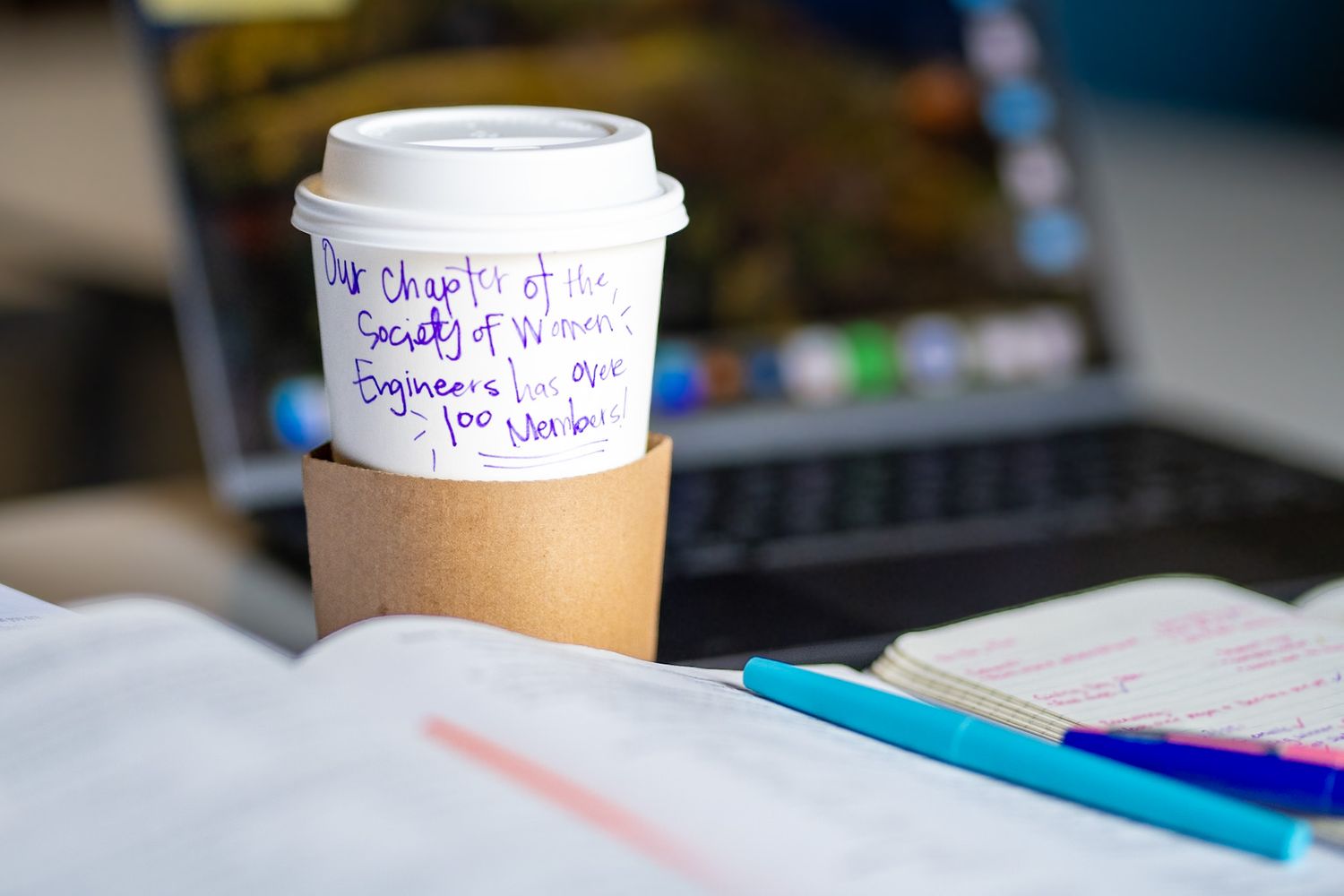 A coffee cup with handwritten Society of Women Engineers message sits on an open notebook near a laptop.