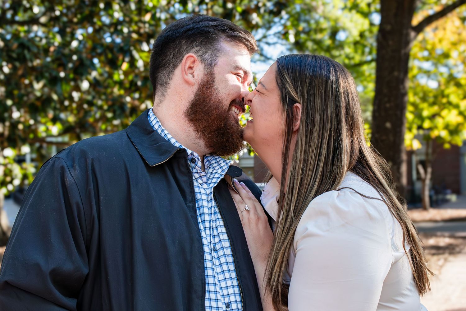 A couple shares a romantic kiss outdoors on a bright autumn day among trees with golden sunlight filtering through.