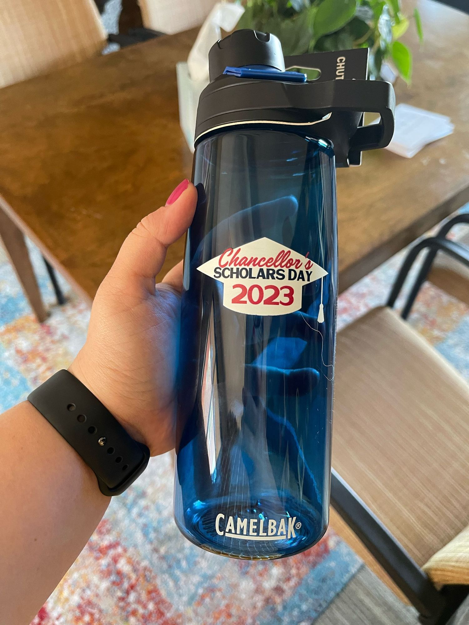 Hand holding a blue CamelBak water bottle with Chancellor's Scholars Day 2023 logo sticker indoors.