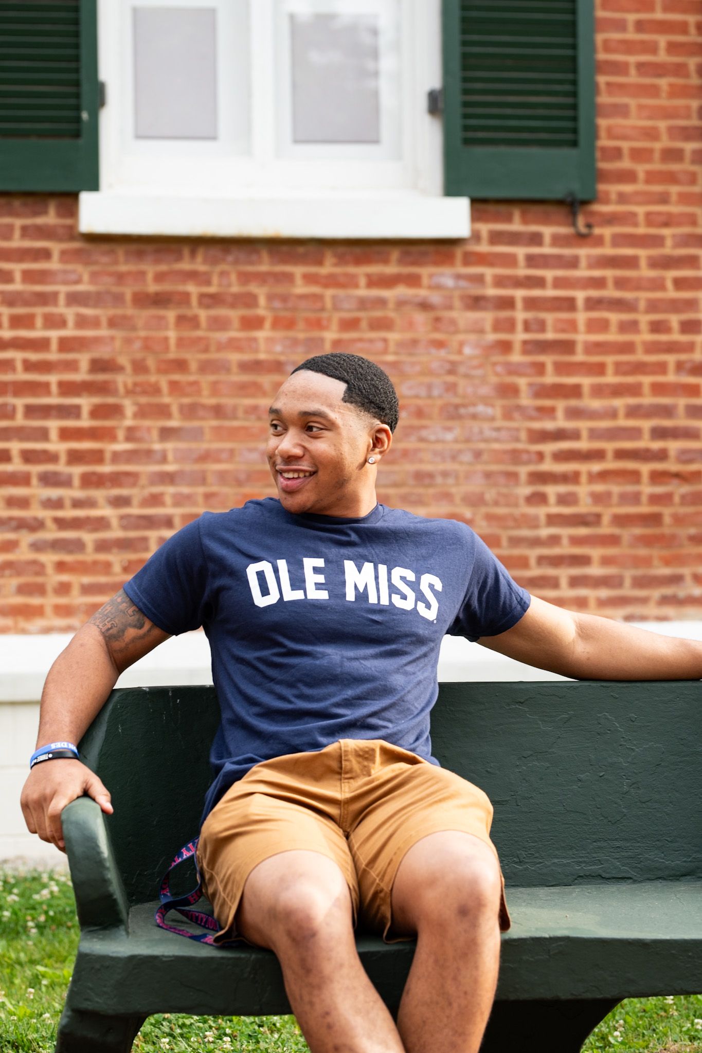 Smiling young man in Ole Miss navy t-shirt sitting relaxed on green bench against brick building.