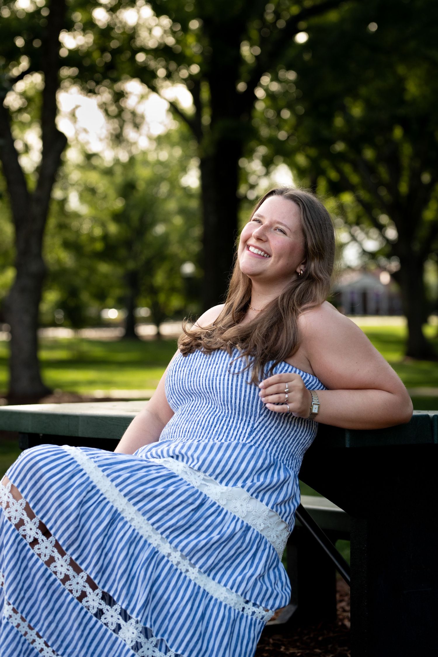 A smiling young woman in a blue striped sundress sits on a park bench, laughing joyfully surrounded by lush green trees.